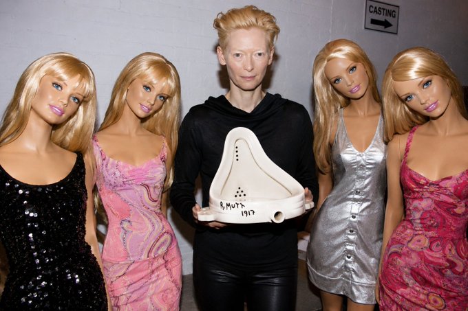 Tilda Swinton poses backstage at a 'CASTING' area between four blonde Barbie dolls, holding a signed white urinal marked 'B. Mutt 1997.'