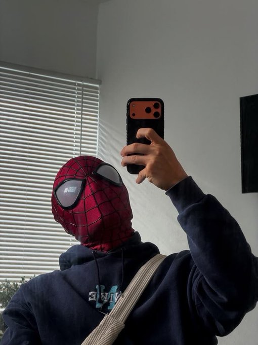 A mirror selfie of a person wearing a dark blue hoodie and a Spider-Man mask, holding up a phone with an orange case.