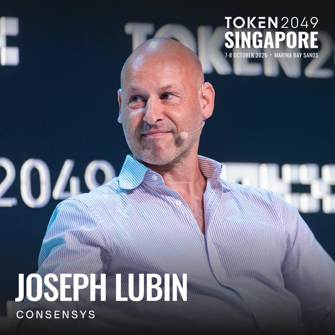 Joseph Lubin, CEO of Consensys and Ethereum co-founder, is shown on stage at TOKEN2049 Singapore (7-8 October, Marina Bay Sands) wearing a striped button-down shirt and headset microphone, illustrating his appearance at the crypto conference featured in the post.