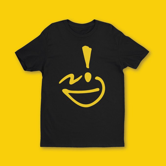 A black t-shirt displayed on a yellow background, featuring a yellow smiley face graphic with an exclamation mark above it.