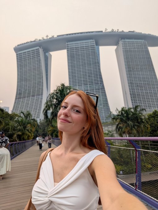 A red-haired woman in a white knotted top takes a selfie with sunglasses perched on her head, standing on a bridge with the iconic Marina Bay Sands hotel looming behind her in Singapore. The image captures the post-Token2049 conference vibe—balancing crypto conference hustle ("too much alpha") with the jet-lagged exhaustion of a Singapore evening, promising a return next year.