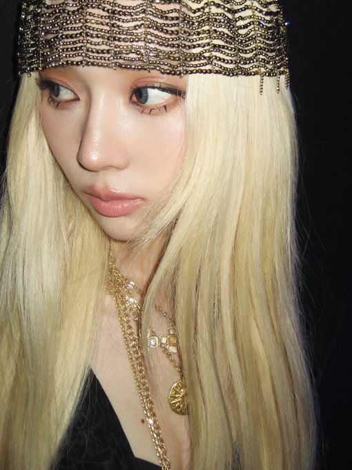 Close-up portrait of a woman with long platinum blonde hair wearing an ornate gold beaded headpiece, layered gold necklaces, and glossy makeup.