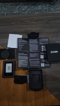 An unboxing layout of EBA branded electronics accessories including documentation, a small black device, a coin-like item, and packaging materials on a wooden surface.