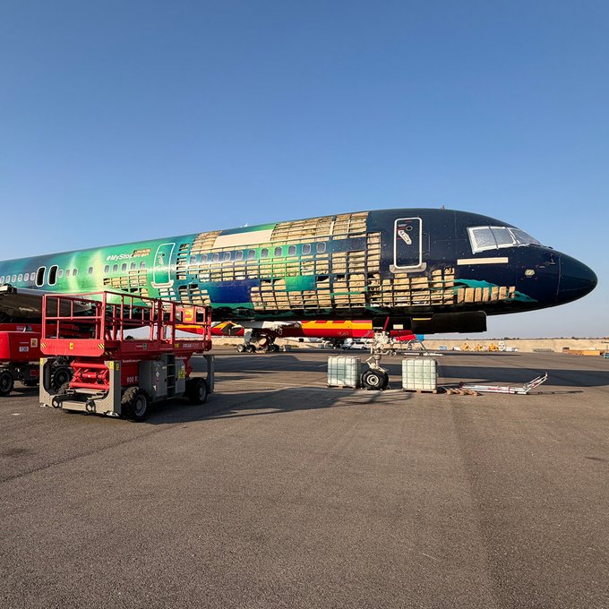 A Boeing 737 aircraft bearing partial "Hekla Aurora" livery sits on a desert tarmac, its fuselage wrapped in protective covering and masking tape during the painting process, with a red scissor lift positioned alongside for workers to access the upper surfaces. The exposed green and dark blue gradient visible on the tail and fuselage reflects the "green aurora" paint scheme the passenger has chosen a stake in.