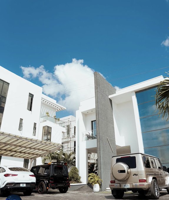 A modern white multi-story building stands under a bright blue sky, with luxury vehicles including a white Mercedes and gold/champagne Mercedes G-Wagon parked outside.