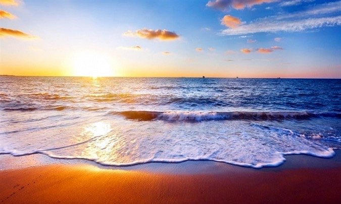 The image shows a beautiful beach scene with a sunset over the ocean, featuring a clear blue sky filled with clouds and the sun setting behind the water. The photo captures the essence of a serene and picturesque moment at the beach.