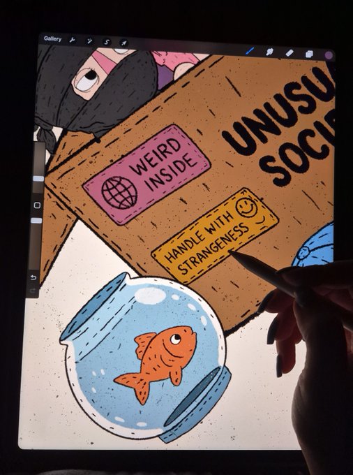 A hand uses a stylus to draw on a tablet, illustrating a cardboard box labeled 'WEIRD INSIDE', 'UNUSUAL SOCIAL', and 'HANDLE WITH STRANGENESS', featuring a fishbowl with a goldfish and a masked character.