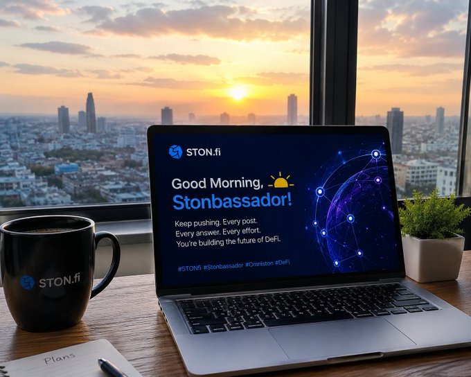 The image shows a laptop computer sitting on top of a wooden desk with a cup of coffee next to it. The screen of the laptop displays a message that reads "Good morning Stonbassadors If you’re helping people understand what’s happening with Omniston and cross-chain swaps, keep going". The background features a cityscape at sunset, which adds an artistic touch to the scene.