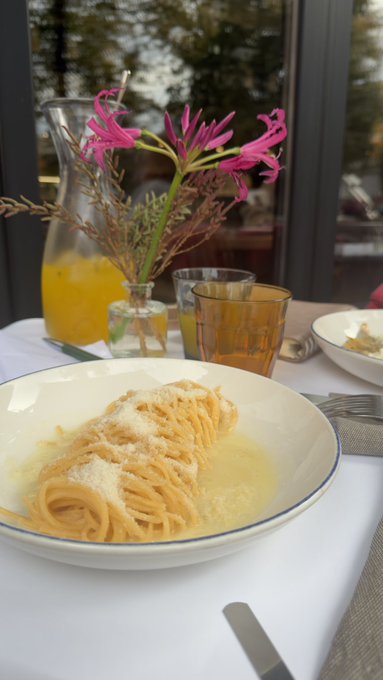 A plate of pasta topped with grated cheese, served on a table with a glass carafe of orange liquid, drinking glasses, and a small vase of pink flowers.