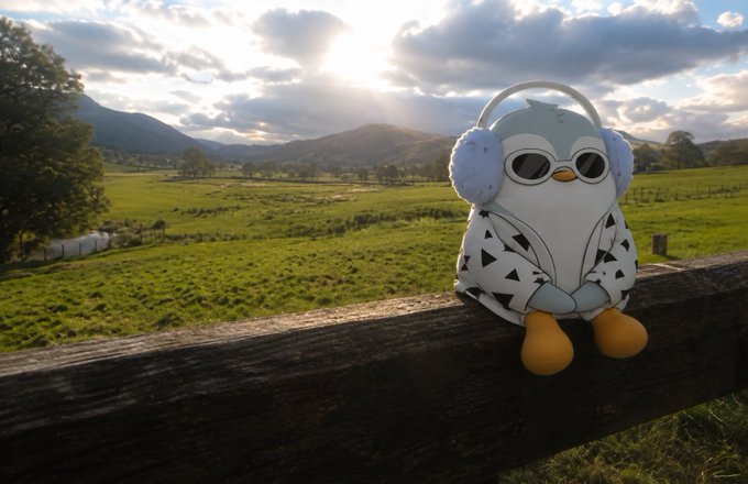 A small plush penguin toy wearing sunglasses and headphones sits on a wooden fence, with a scenic green pasture, mountains, and cloudy sky at sunset in the background.