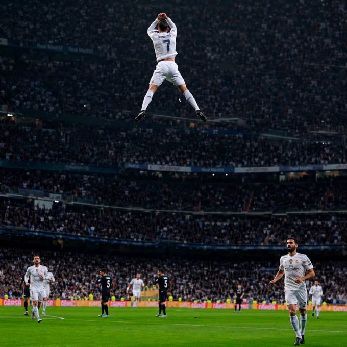 A soccer player wearing a white Real Madrid jersey with the number 7 and 'RONALDO' on the back leaps high in the air in celebration at a packed stadium, while teammates look on.