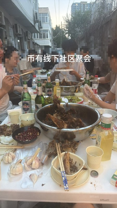 A group of people gather around an outdoor table piled with home-cooked dishes, including a large pot of braised fish, garlic, peanuts, and various beers, with the Chinese text "币有线下社区聚会" (meaning "Coin has offline community gathering") overlaid on the image. The casual street-side feast humorously matches the post's caption about a "down-to-earth" crypto community party.
