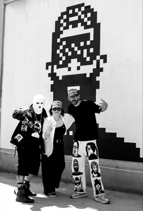 Three people in streetwear, including hats with "NORMIE" text, pose in front of a large pixelated graphic, visually representing the "Normies" mentioned in the accompanying post.