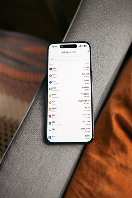 A smartphone rests on a gray cushioned surface next to an orange fabric, displaying a cryptocurrency prices app showing tickers like ADA (+1.65%), AVAX (-2.51%), AXS, BCH, BNB, BTC (+1.60%), DOGE (+4.10%), DOT, ETC (+2.01%), ETH (+0.29%), and FIVE. The market data on screen visually reinforces the post's message about weathering crypto volatility with long-term conviction.