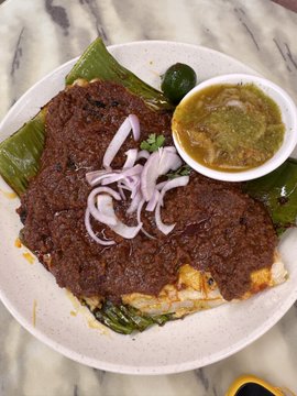 A plate of food featuring a dark, rich curry or stew garnished with sliced red onions, served on a banana leaf alongside a small bowl of green chutney.