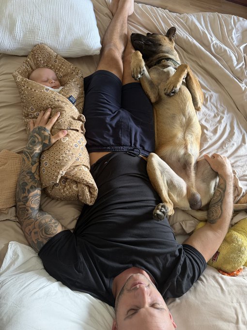 A tattooed father lies asleep on his back in bed, cradling a swaddled newborn beside him while his tan dog sleeps belly-up on his chest. The image captures a peaceful family nap scene, fitting the caption "Shhhh do not wake my babies."