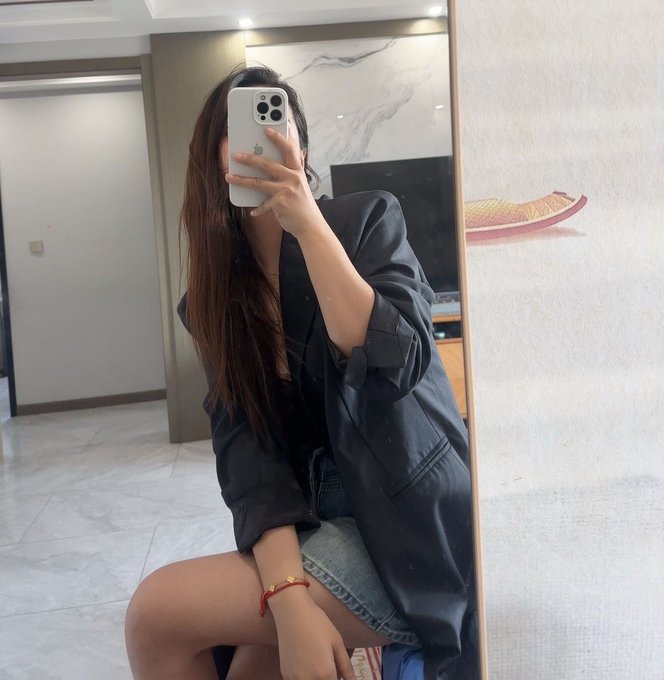 A young woman with long dark hair takes a mirror selfie with an iPhone, wearing an oversized dark blazer, a denim skirt, and a red string bracelet.