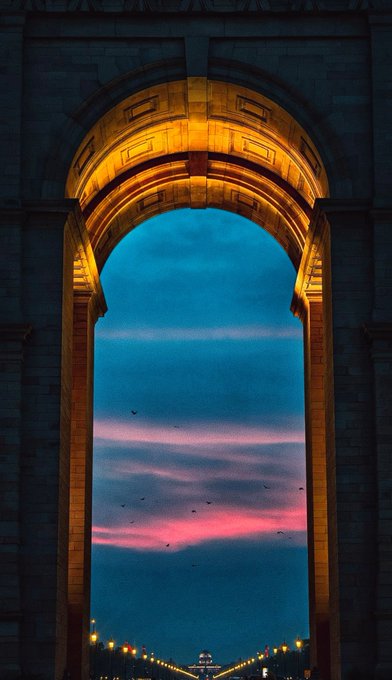 A grand stone archway glows with golden light at twilight, framing a vibrant sunset sky streaked in blue and rose hues, with distant lights and a small monument visible far below. The image perfectly embodies the "Heaven Portal" theme—capturing how the illuminated arch acts as a luminous doorway, through which the evening sky unfolds like a breathtaking view into a gilded, otherworldly realm.
