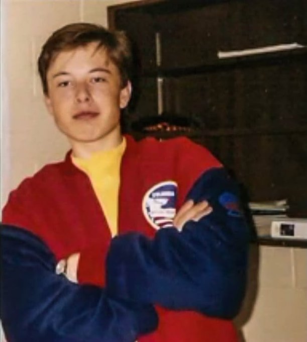A young man stands with arms crossed in front of a dark microwave, wearing a red and blue jacket featuring a France Football Federation logo over a yellow shirt.