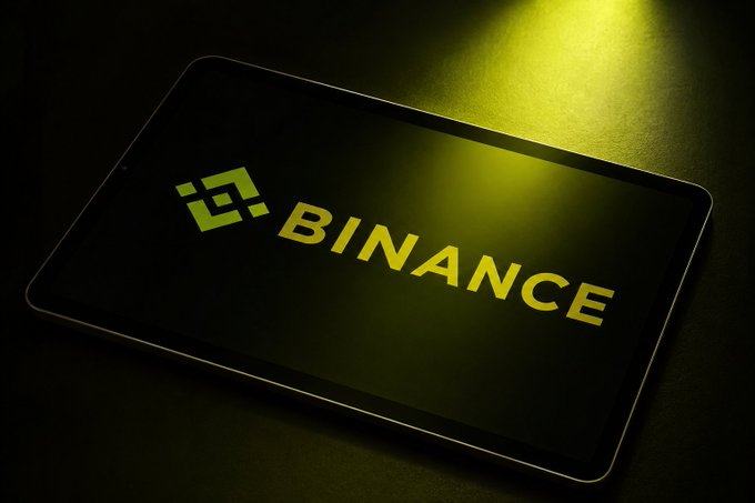A smartphone displays the Binance cryptocurrency exchange logo in yellow-green on a dark screen, illuminated by a warm light.
