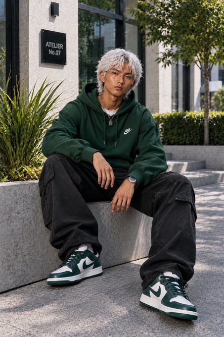 A young man with white hair sits on a stone ledge outdoors wearing a dark green Nike hoodie, black cargo pants, and green and white sneakers. A sign behind him reads "ATELIER No.07".