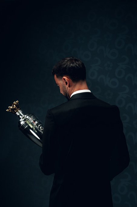 A man in a dark suit stands with his back to the camera, holding a silver trophy with gold figures on top against a dark background.