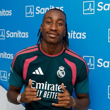 A young male soccer player with braided hair poses pointing at the Real Madrid crest on his new dark green and pink Adidas jersey with 'Emirates Fly Better' sponsor, against a blue Sanitas backdrop.