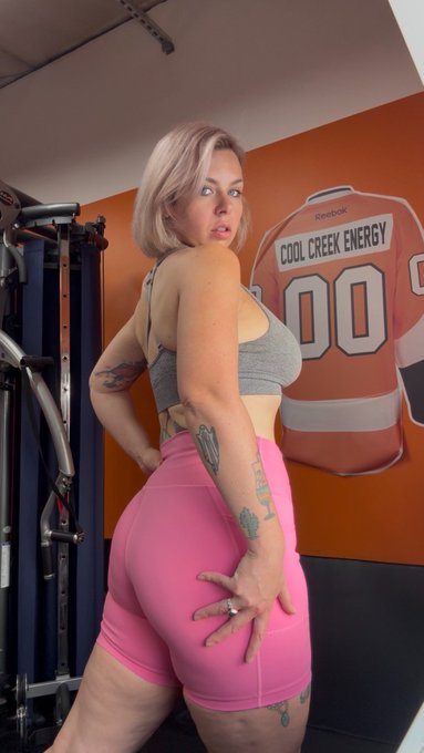 A woman with pink hair poses in a gym wearing a gray sports bra and pink shorts, with a 'COOL CREEK ENERGY' jersey visible on the wall behind her.