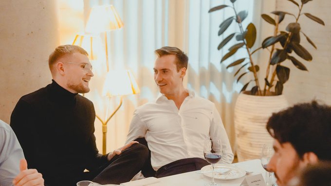 Two men in business attire smiling and conversing at what appears to be a dinner event, with one in a black turtleneck and glasses, the other in a white shirt, surrounded by warm lighting and a potted plant.