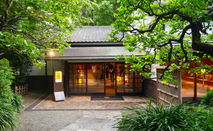 A serene view of a Japanese-style building entrance with a tiled roof, surrounded by lush green trees, with warm light glowing through the glass doors.