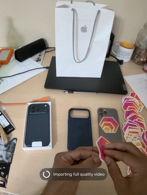 A person unboxing an iPhone, with the phone case and stickers visible on a desk alongside an Apple shopping bag on top of a laptop, with a message indicating 'Importing full quality video'.