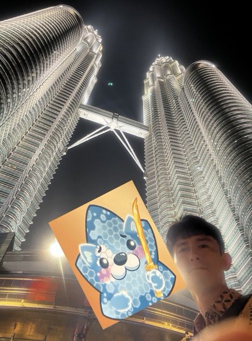 A person holds up a photo of a cute blue shark cartoon mascot in front of the illuminated Petronas Twin Towers at night.