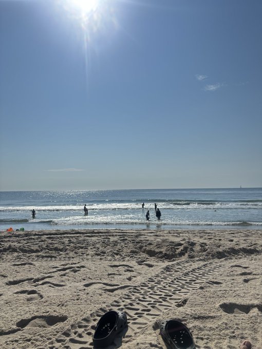 A sunny beach scene with blue skies, gentle waves, a few people in the water, and a pair of shoes left on the sandy shore in the foreground.