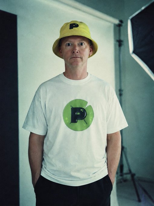 A man wearing a yellow bucket hat with a black 'P' and a white t-shirt featuring a green circle with a black 'P' stands in front of a photography backdrop with lighting equipment.