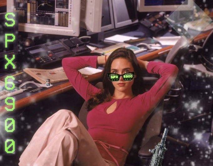 A woman in a pink top relaxes in a chair within a retro trading setup, wearing pixelated sunglasses displaying 'SPX 6900', with 'SPX 6900' glowing in green text on the left side.