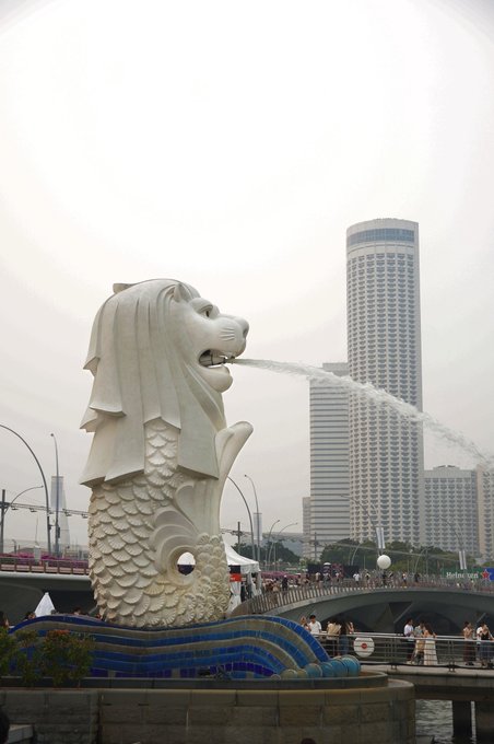 A photo of the Merlion statue in Singapore spouting water, with the iconic Marina Bay skyscrapers and a cloudy day in the background.