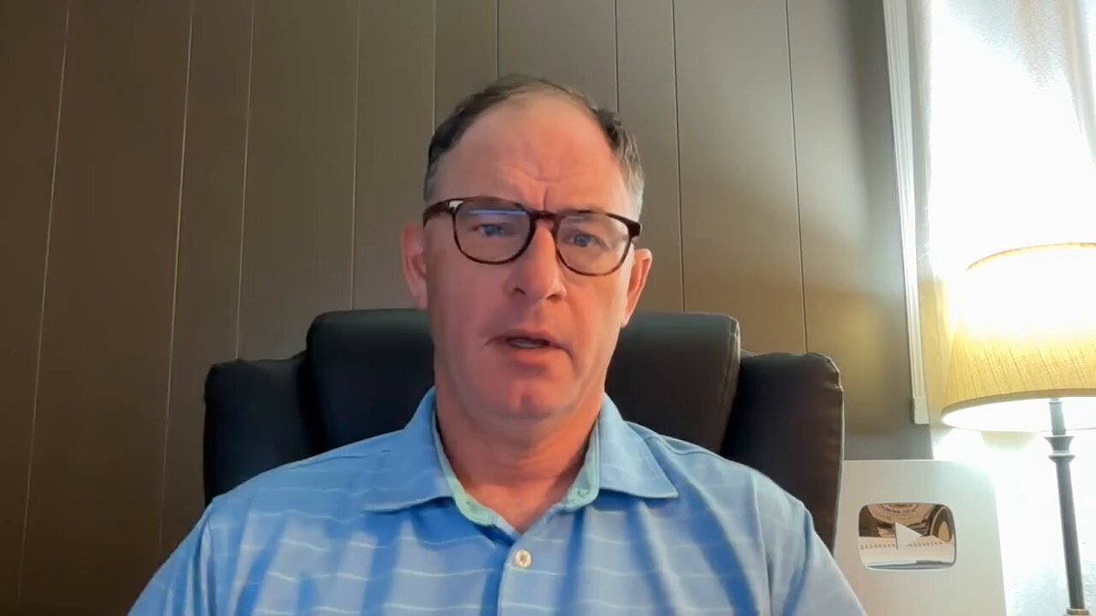 The image shows a man wearing glasses sitting in an office chair, looking at the camera with a serious expression. He appears to be engaged in a video call or participating in a live stream event. The background features a wall with vertical wooden panels, which adds to the professional atmosphere of the setting.