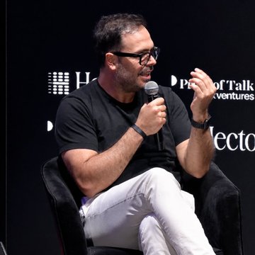 The image features a man sitting in a chair, holding a microphone and talking into it. He appears to be wearing glasses and has short hair. The background of the image shows a black wall with white text that reads "BREAKING: Costas Chalkias Co-founder of Mysten Labs says RWAs are becoming more liquid and will eventually merge with DeFi." This suggests that the man is discussing the concept of tokenized assets, which could be related to the emerging technology called DeFi.

The image also includes a chair in front of the man, indicating that he might be at an event or gathering where people sit while listening to speakers like him. The presence of the chair and the microphone suggests that this is likely taking place during a talk or presentation on the topic of tokenized assets and their potential impact on the financial industry.