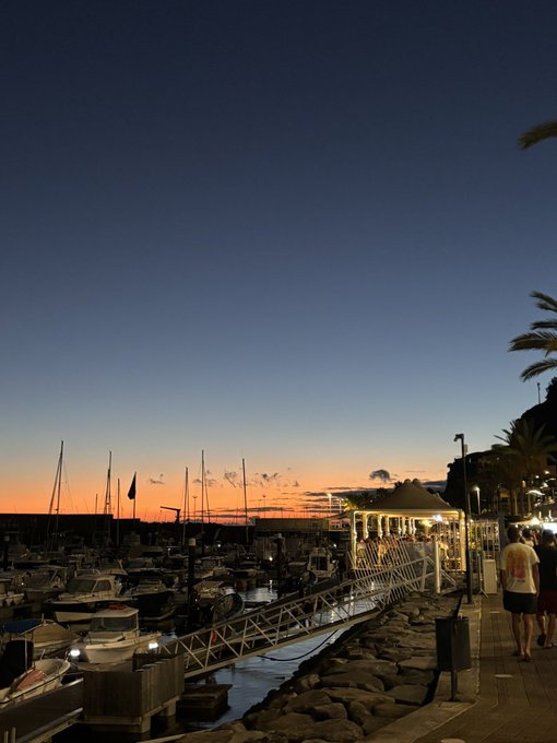 A scenic sunset harbor with sailboats docked and a lit gazebo along the waterfront promenade, where a few people are walking. The image doesn't directly relate to the "Gm fam / Reply with SOL addy for a surprise" message, but the calm, inviting seaside setting could fit a relaxed social media greeting vibe.