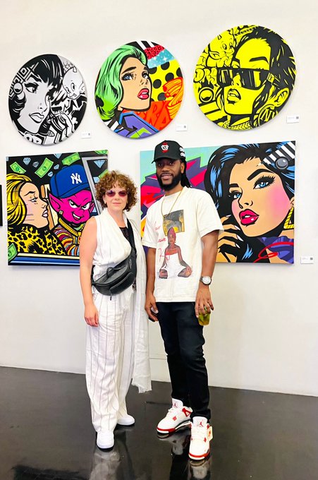 Two people pose in front of pop-art style paintings featuring stylized faces in vibrant colors and bold outlines at what appears to be an art gallery.