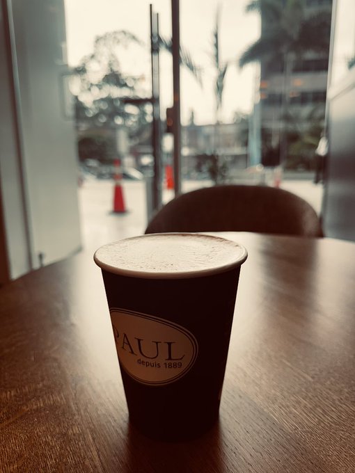 A cup of coffee from PAUL (depuis 1889) sits on a wooden table, with an outdoor street scene featuring orange traffic cones visible through the window behind it.