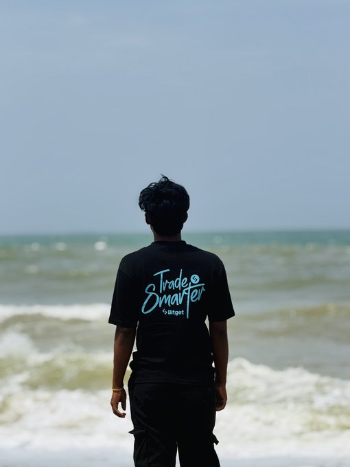A photograph of a person viewed from behind standing on a beach, wearing a black t-shirt with the text 'Trade Smarter by Bitget' printed in cyan.