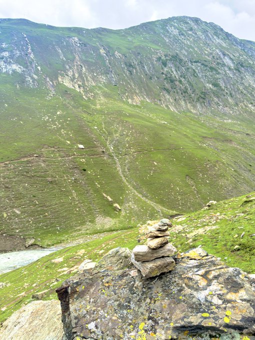 A scenic mountain landscape featuring a small stack of stones (cairn) balanced on a rocky outcrop in the foreground, with steep grassy slopes rising into rugged mountains under a partly cloudy sky.