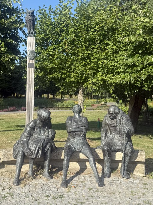 An outdoor bronze sculpture depicting three seated figures on a stone bench, with a tall wooden post topped by an owl statue and a face medallion behind them, set in a park with green trees.