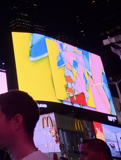 A nighttime street photograph of a brightly lit digital billboard displaying colorful abstract art, positioned above a McDonald's restaurant, with the back of a person's head visible in the foreground.