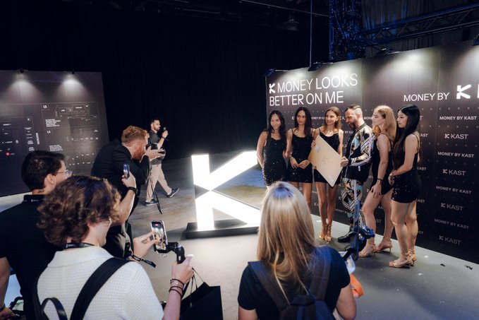 A promotional event for 'KAST' where models pose under a sign reading 'MONEY LOOKS BETTER ON ME MONEY BY KAST,' while photographers and videographers capture the scene around a large illuminated 'K' logo.