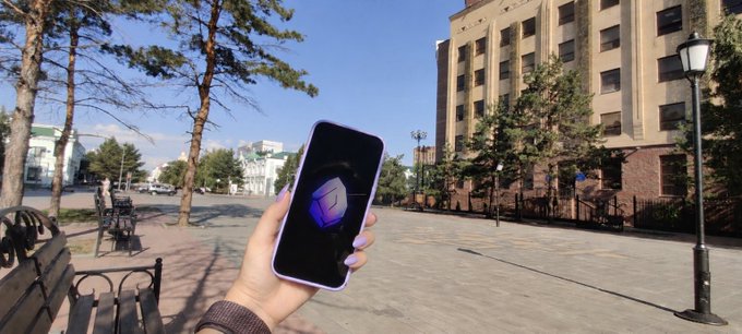 A hand holds up a smartphone displaying a purple logo or graphic in front of a sunny outdoor plaza with trees, benches, and a multi-story building in the background.