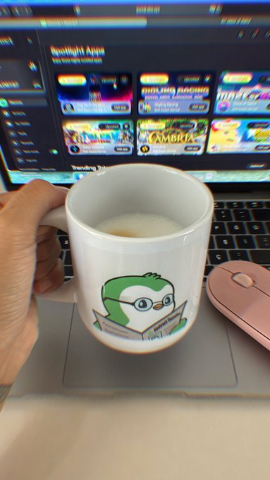 A hand holds a white Duolingo owl mug containing a beverage in front of a laptop displaying the Duolingo app and a pink computer mouse.
