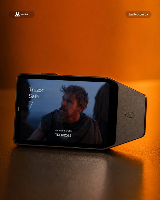 A Trezor Safe 7 hardware wallet sits on an orange-lit surface, its screen displaying a still of a bearded man from "The Odyssey" alongside the text "Trezor Safe 7 — secured with TROPIC01," playfully framing the crypto device as a portable cinema for watching the film on the go.