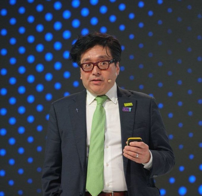 A man in a dark suit, white shirt, and green tie stands on a stage with a blue polka-dot background, wearing glasses and a pin that reads 'DO MORE FUN'.